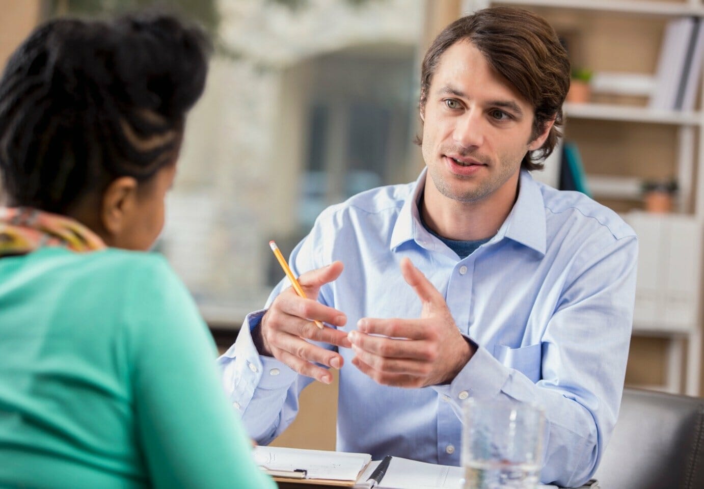 A man in a blue collared shirt holding a pencil in his right hand is speaking to a woman in a green shirt who is sitting across from him at a table.